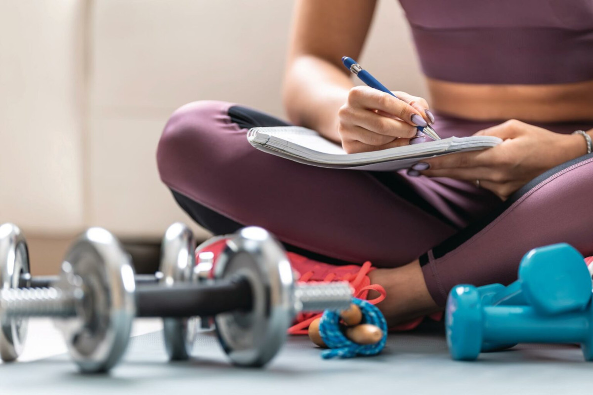 A woman in exercise gear is sat on the floor making notes, surrounded by weights and a skipping rope