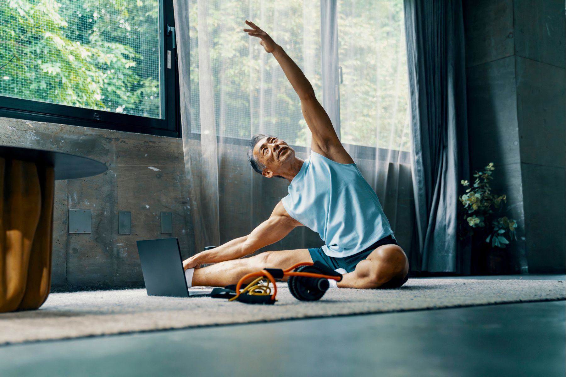 An older man does yoga inside, sat on a mat
