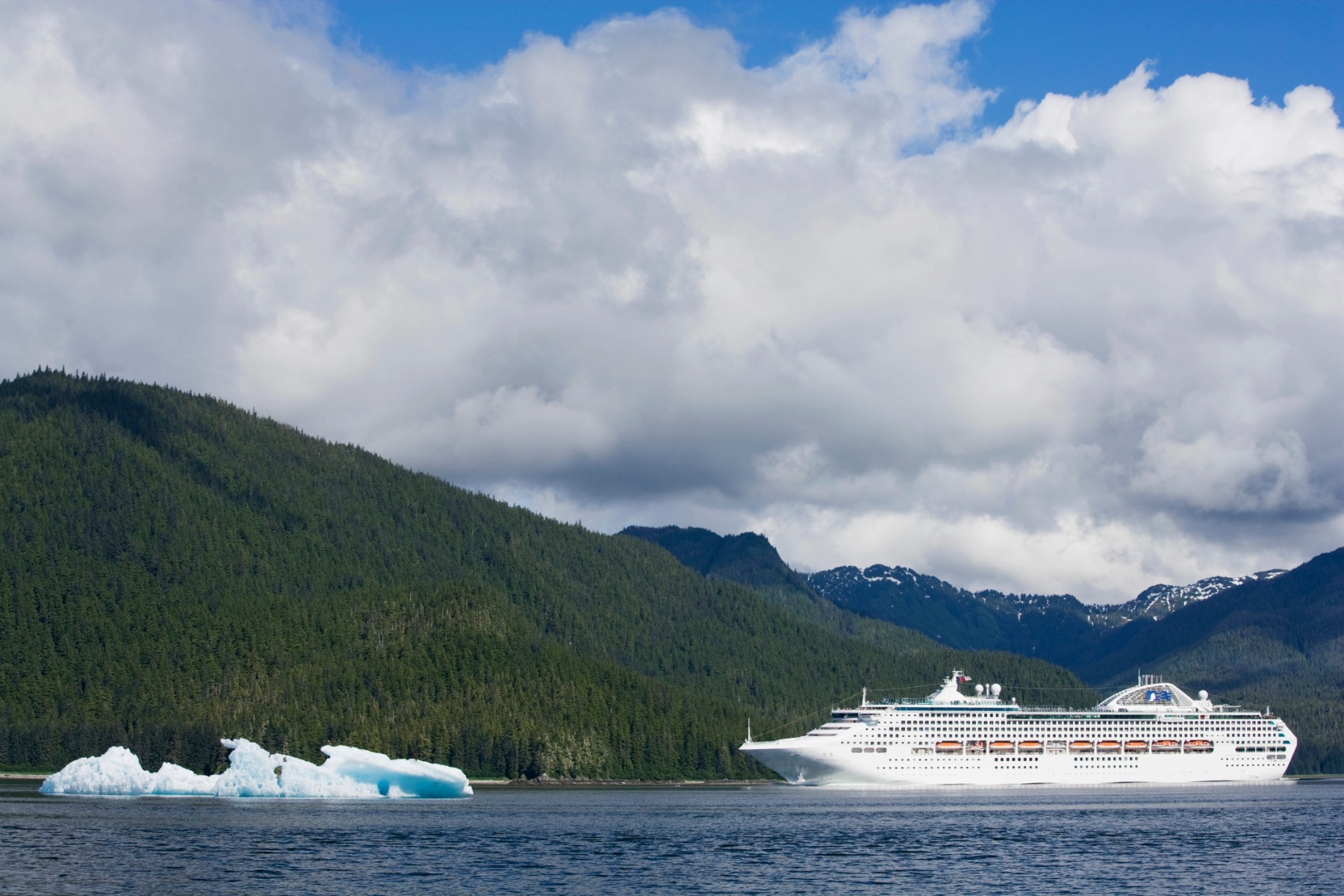 A large white cruise ship sails past lush green mountains and icy water
