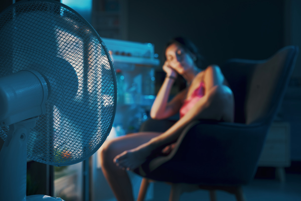 Exhausted woman suffering from the heat during the summer heatwave, she is sitting in front of the open fridge and cooling herself with a fan