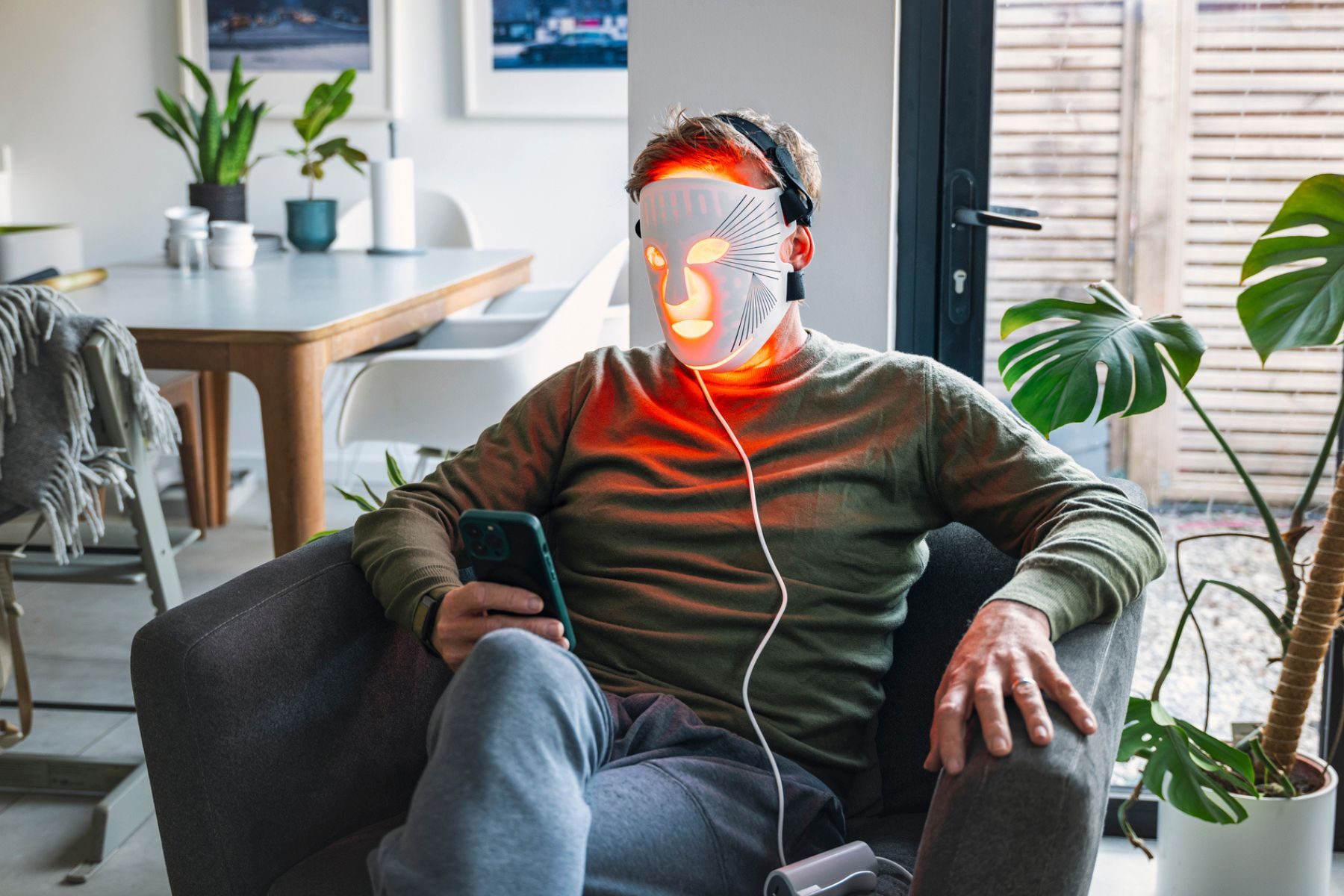 A man sits on an armchair while wearing a red light mask and scrolling on his phone