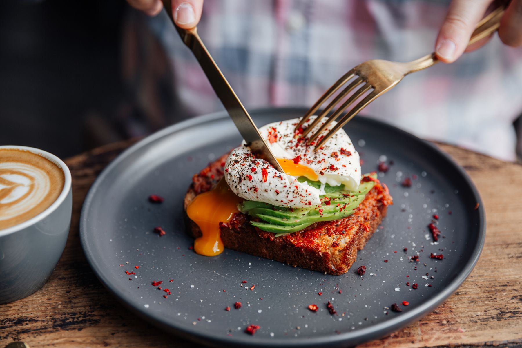 A person uses a knife and fork to break into a poached egg, on top of avocado on toast