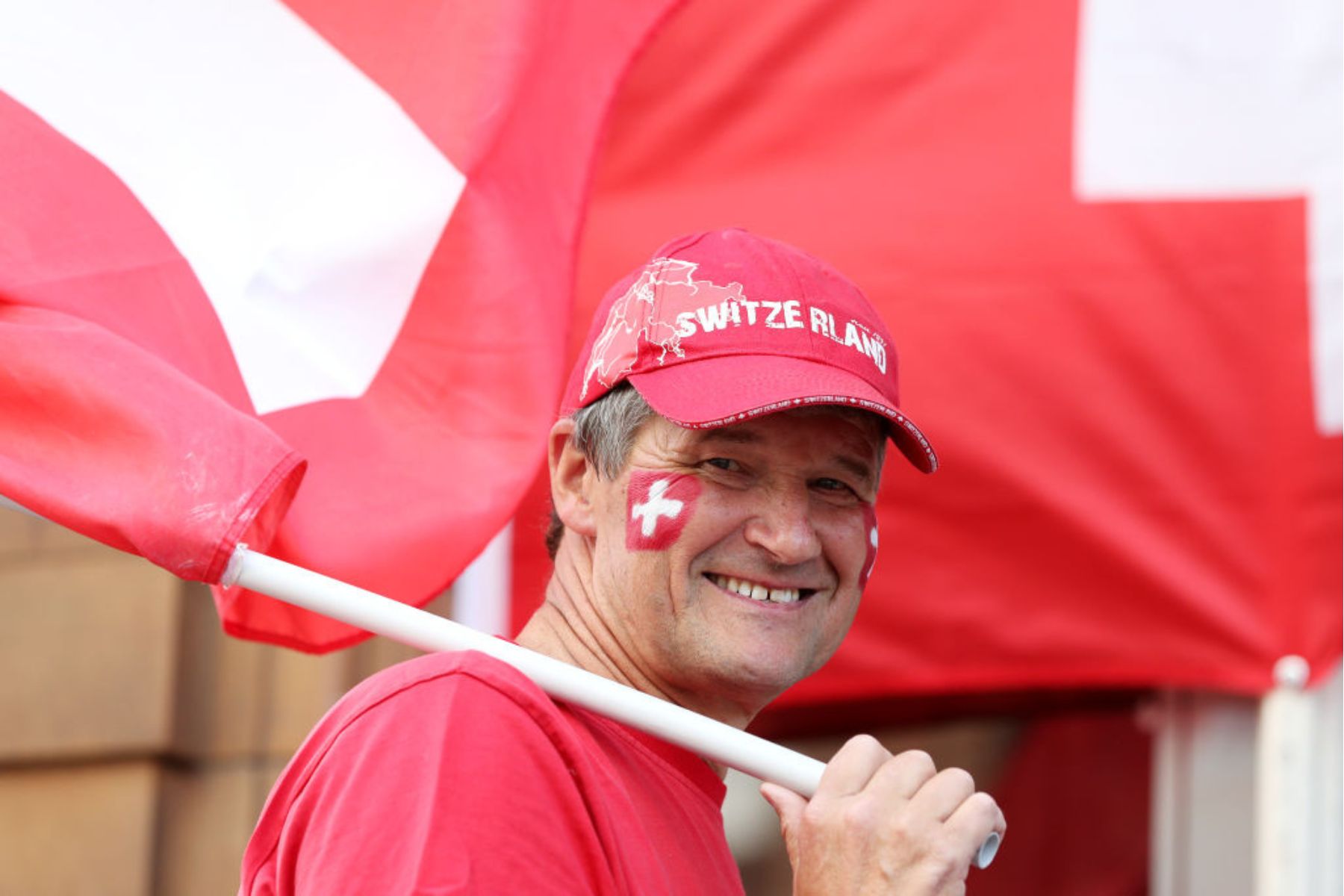 A man holding a Swiss flag, wearing a red Switzerland hat, with face paint of the Swiss flag on his cheek