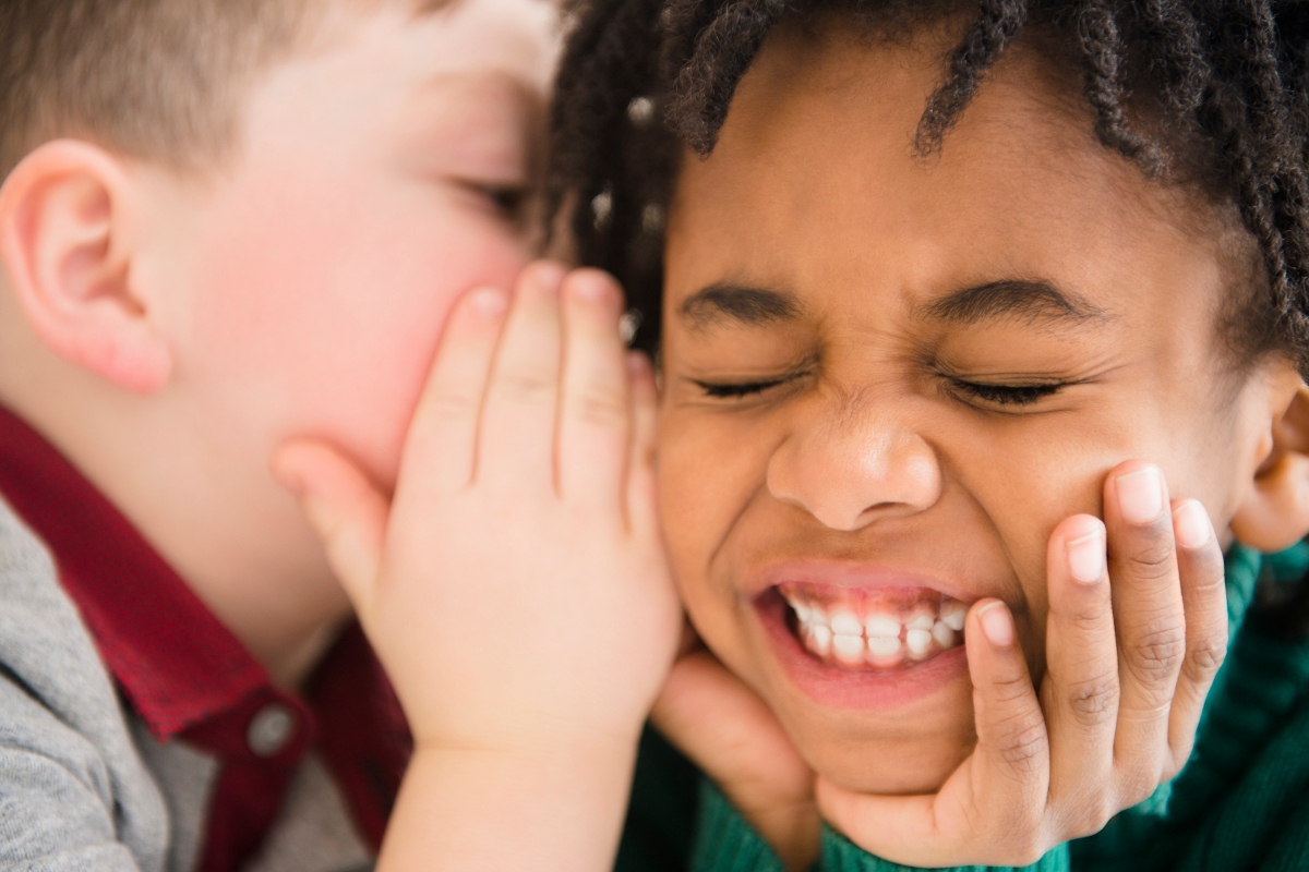 A close-up of two children whispering and laughing
