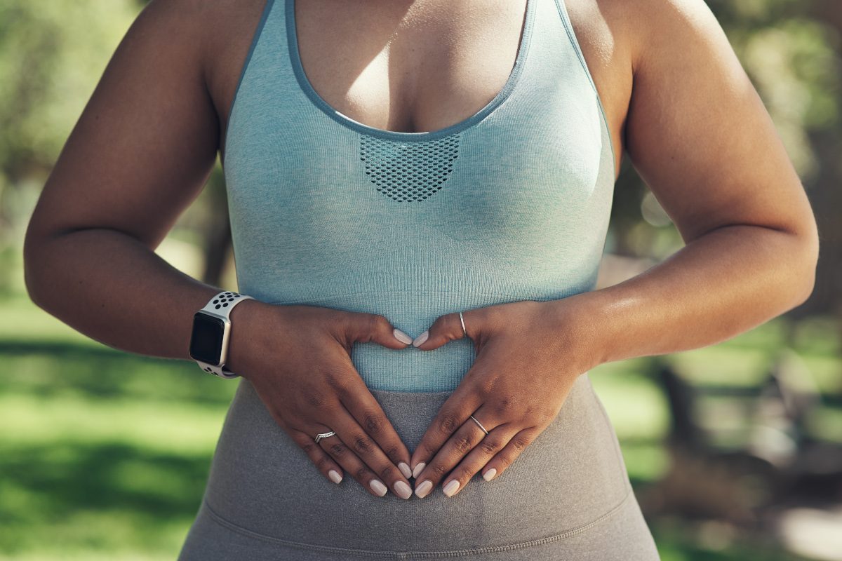 Shot of a woman making a heart symbol with her hands over her gut. She is wearing workout gear and is outside