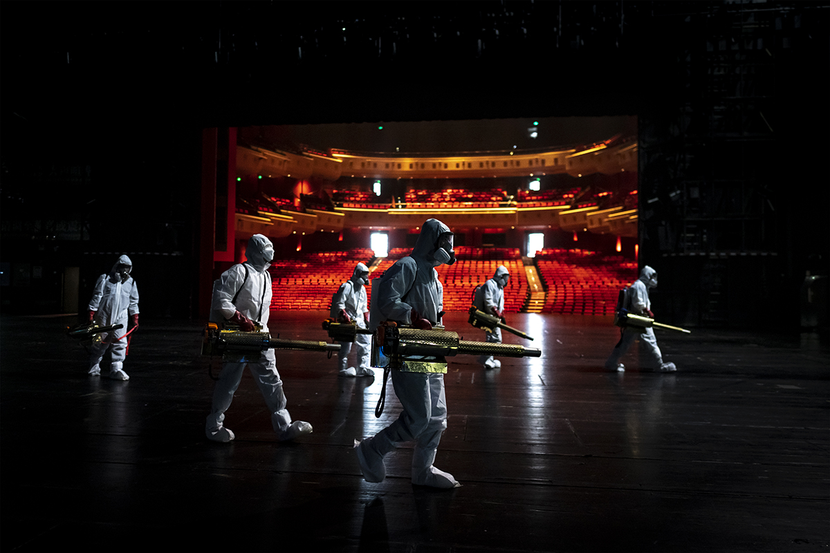 People In hazmat suits walk around on stage of theatre.