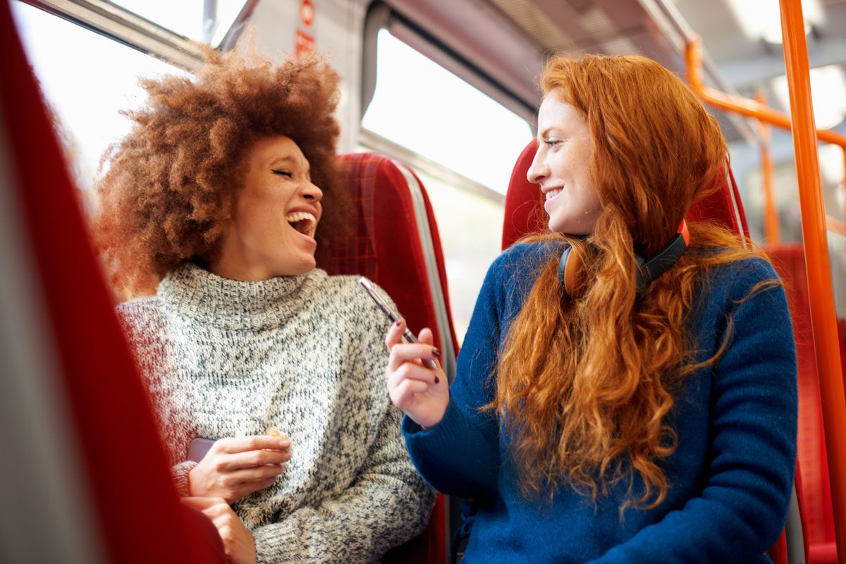 Two friends are sitting on a bus, looking at each other and laughing.