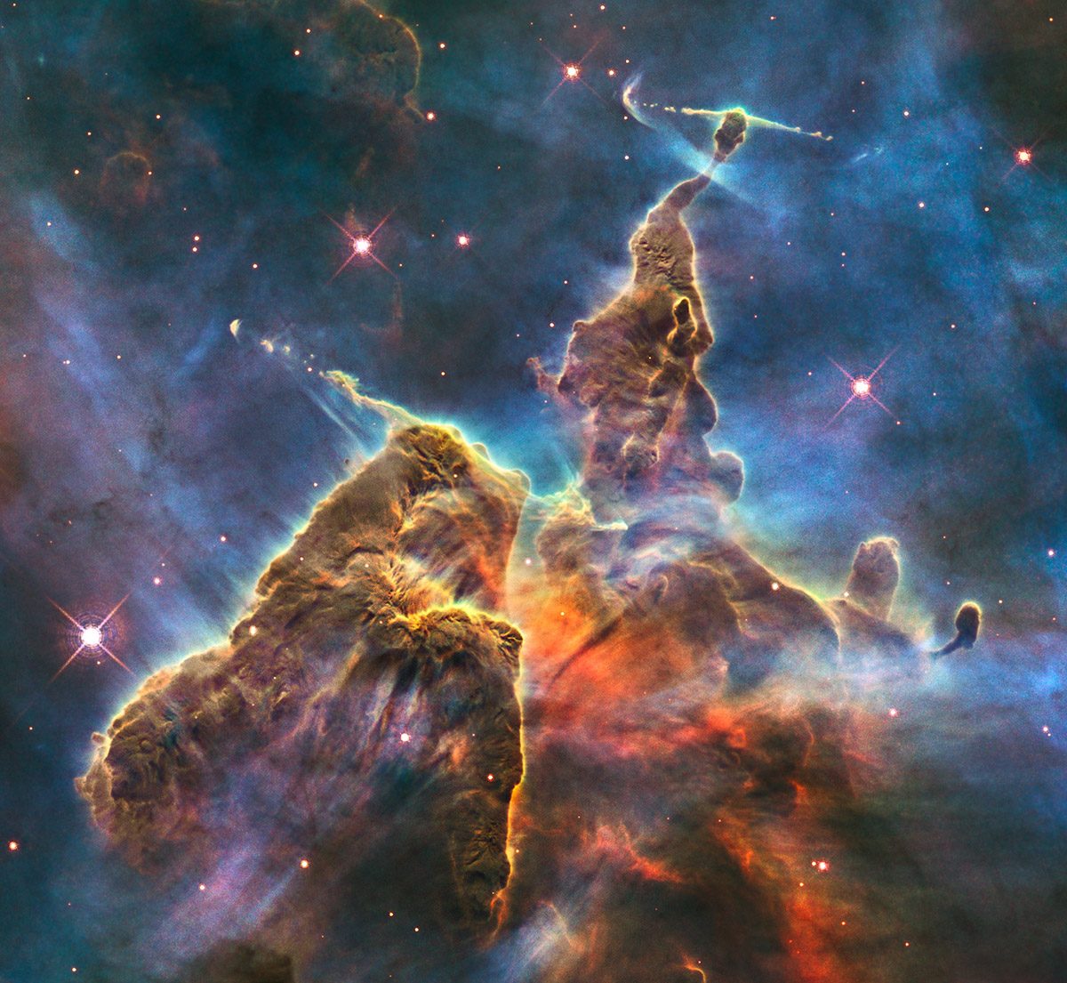 Hubble image of a tall, mountain‑like column of gas and dust with rough, sculpted edges glowing in blue and orange light, surrounded by stars in deep space.
