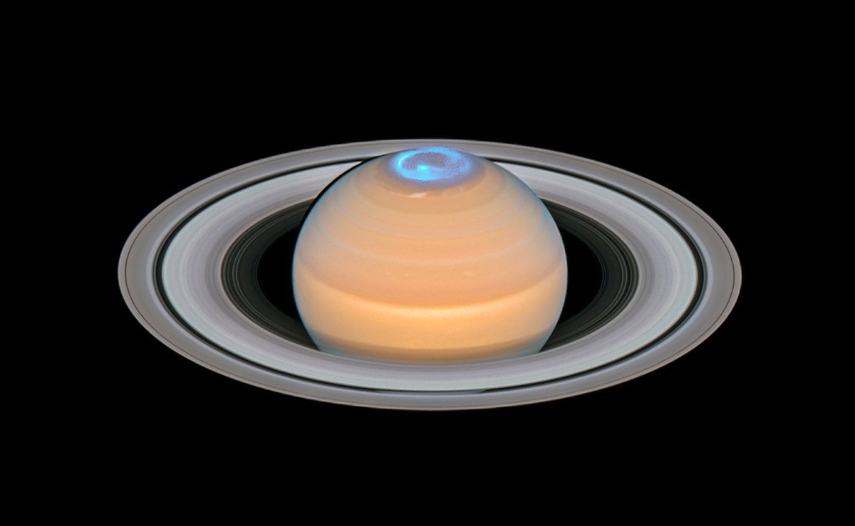 A satelite image of saturn in the centre of the frame in a beige colour with white rings and a blue glow on the top.