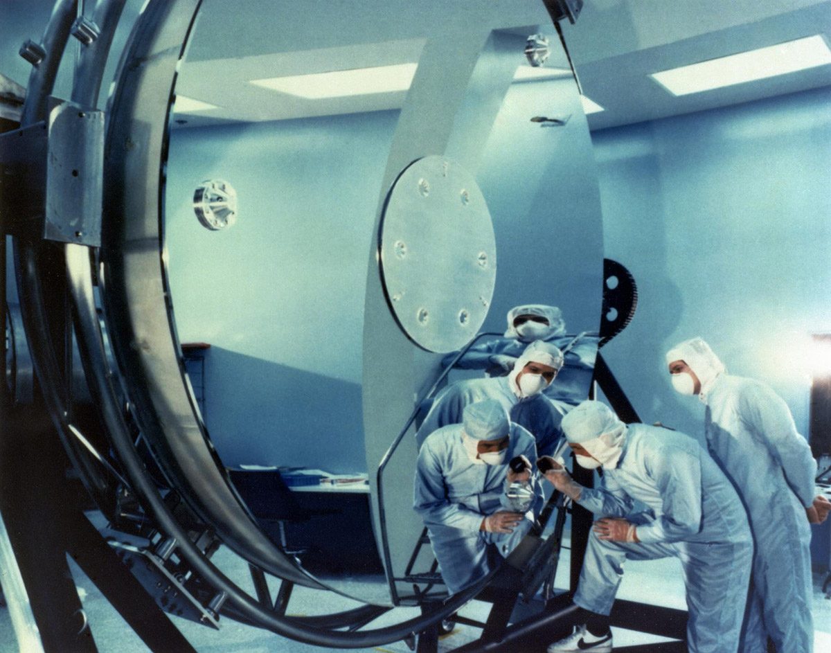 Several technicians in protective suits examining a large reflective cylindrical space instrument inside a clean room.