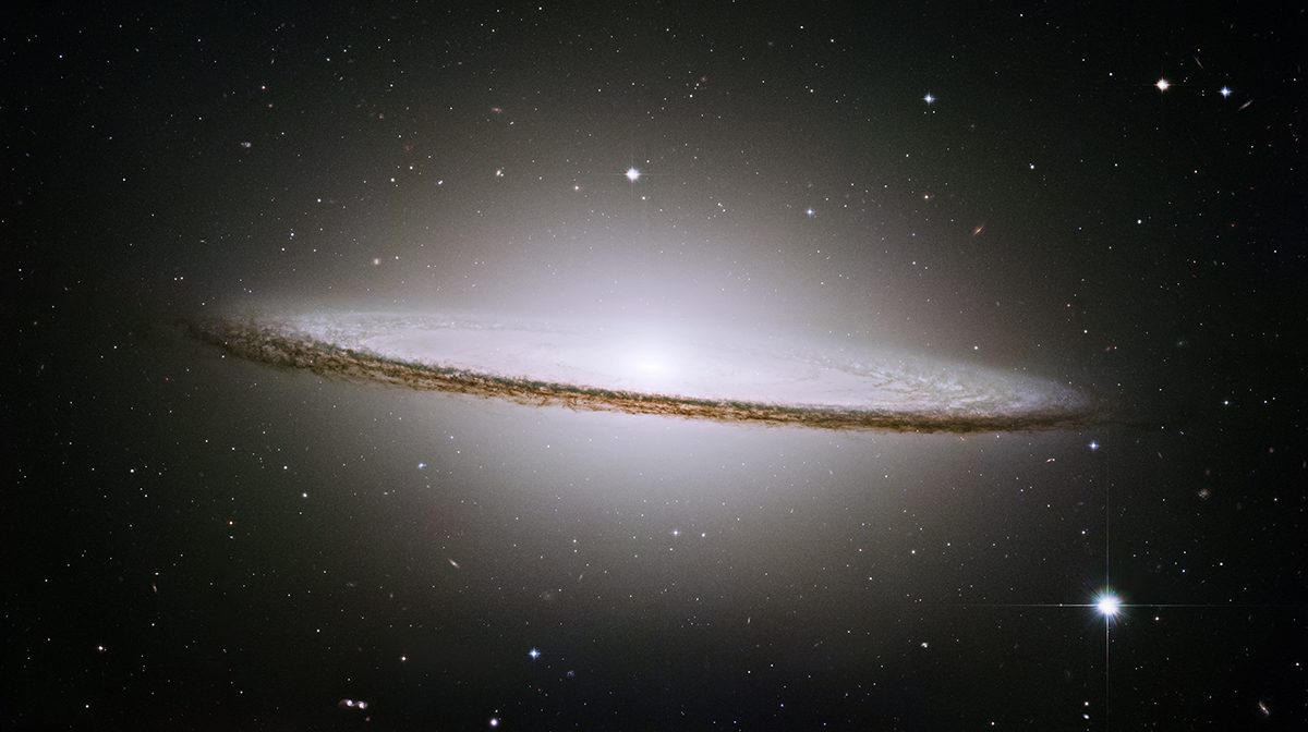 An edge‑on galaxy with a bright central bulge and a dark dust band forming a wide, flat disc against a black background of space.