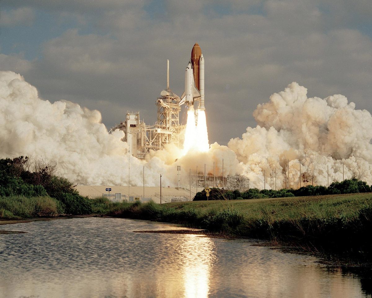 A space shuttle launches from the centre of the image, surrounded by plumes of white smoke and a river leading to the take off site from the foreground.