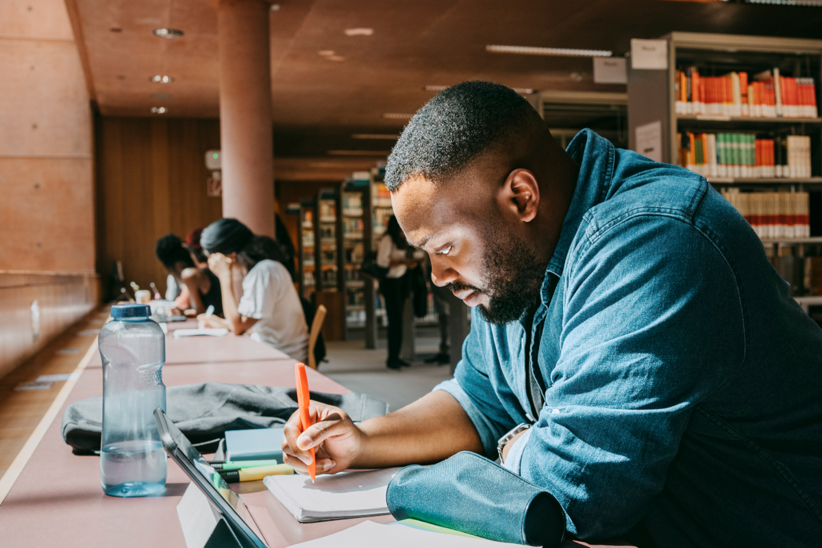 Student writing in a notebook while sitting with tablet PC in a library.