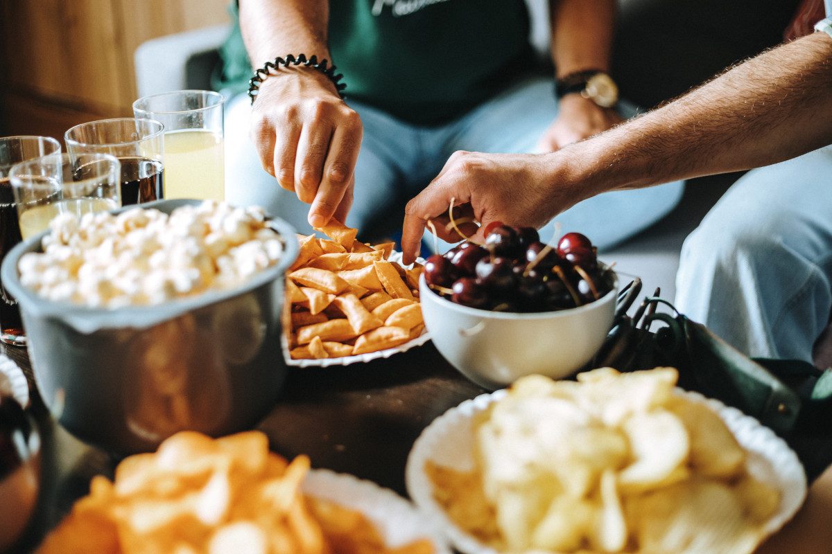 Picture of hands of unrecognizable young people sitting on couch in living room having meal together, reaching for plates with snacks such as cherries, popcorn and potato crisps