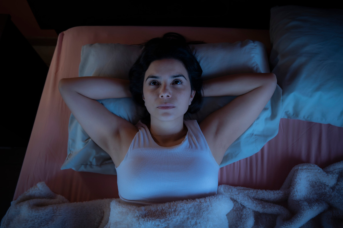 Young woman lying awake in bed at night with hands behind head, staring at the ceiling in a dark bedroom