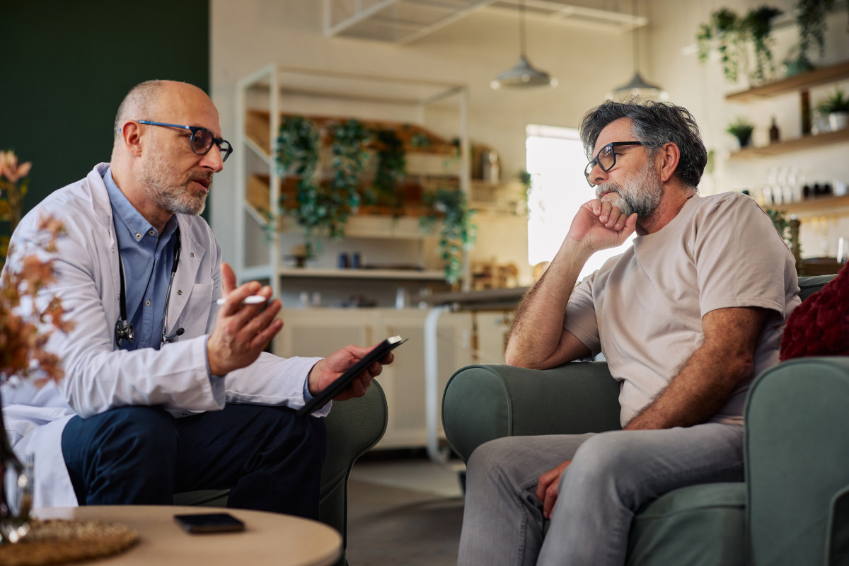 A doctor in a white coat listens attentively to a senior male patient during a private home visit consultation in a warm, plant-filled living room