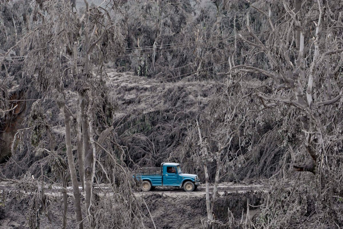 A single blue pickup truck drives along a road through an ash-covered devastated landscape