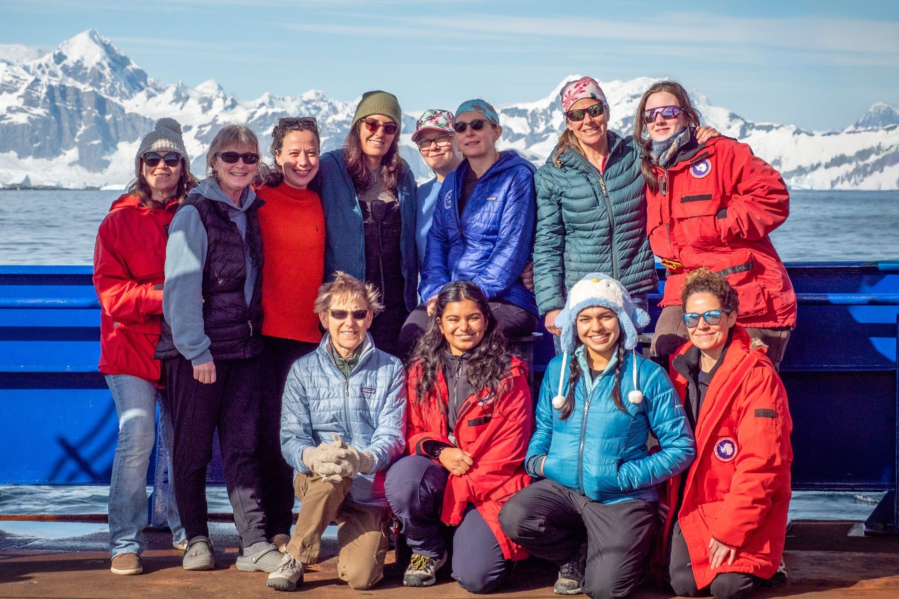 A group of people wearing blue and red stand on a boat, with snowy mountains behind them