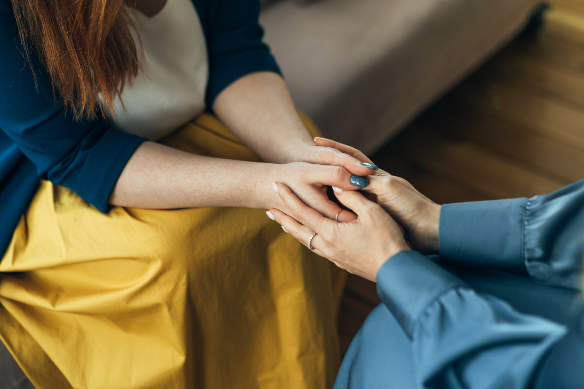 Two women sitting in chairs and talking, focus on their hands holding each other