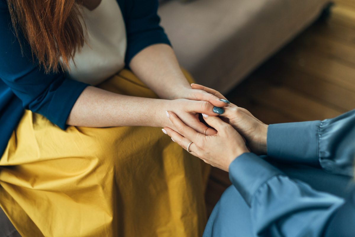 Two women sitting in chairs and talking, focus on their hands holding each other