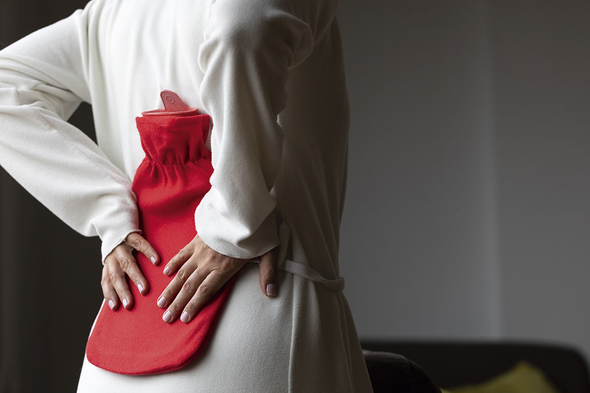 A woman holds a hot water bottle against her back.
