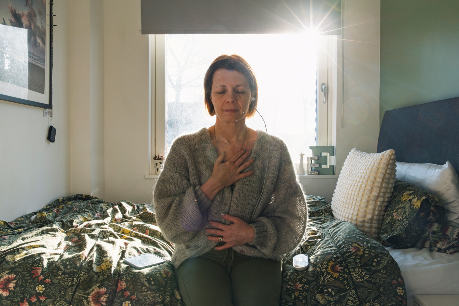 A woman sat on her bed with her eyes closed. One hand is placed on her chest, the other on her stomach