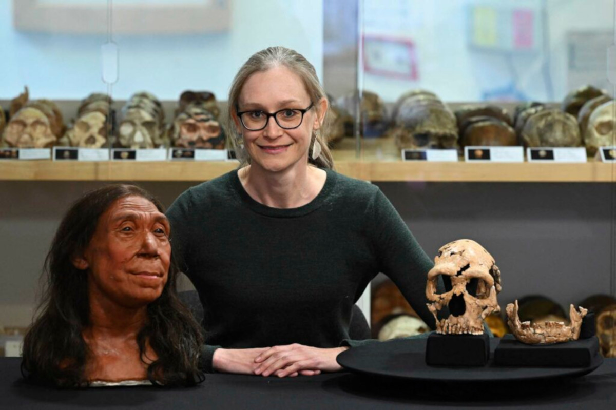 Photo of Dr Emma Pomeroy sat a table with a replica face and skull of ancient humans, rows of skulls are displayed in the background