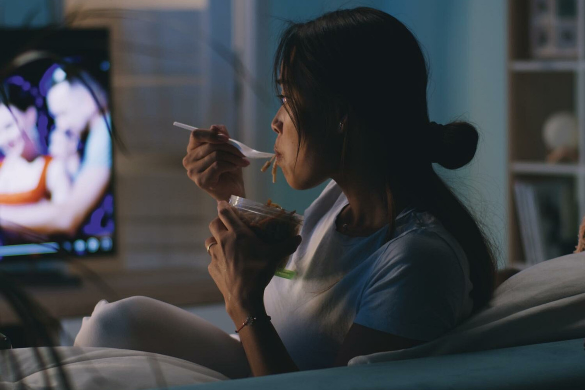 A woman sat on a sofa eating noodles from a microwave dish watching the TV