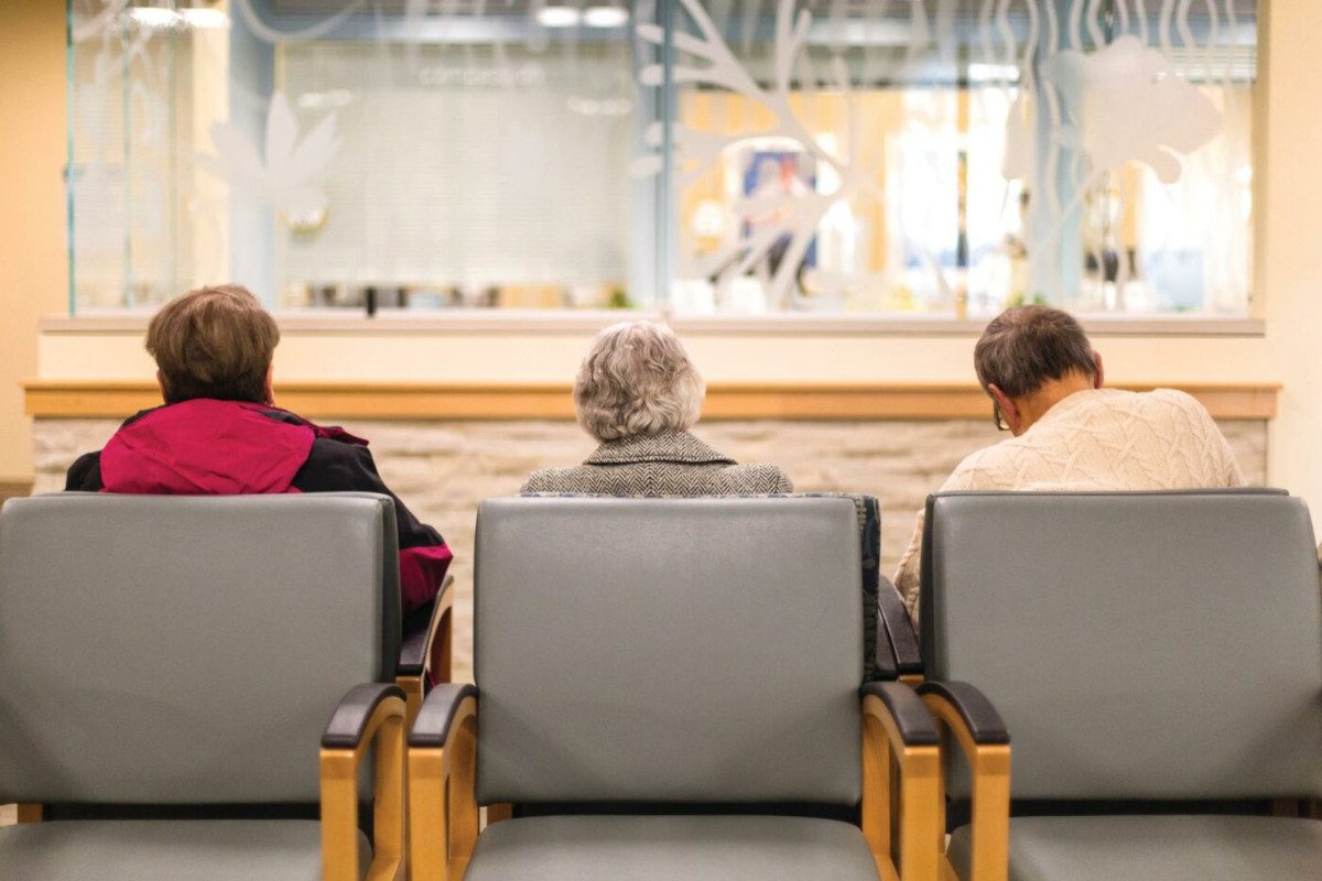 Photo of three elderly people sat in a waiting room of a doctor's surgery. Their backs are to the camera