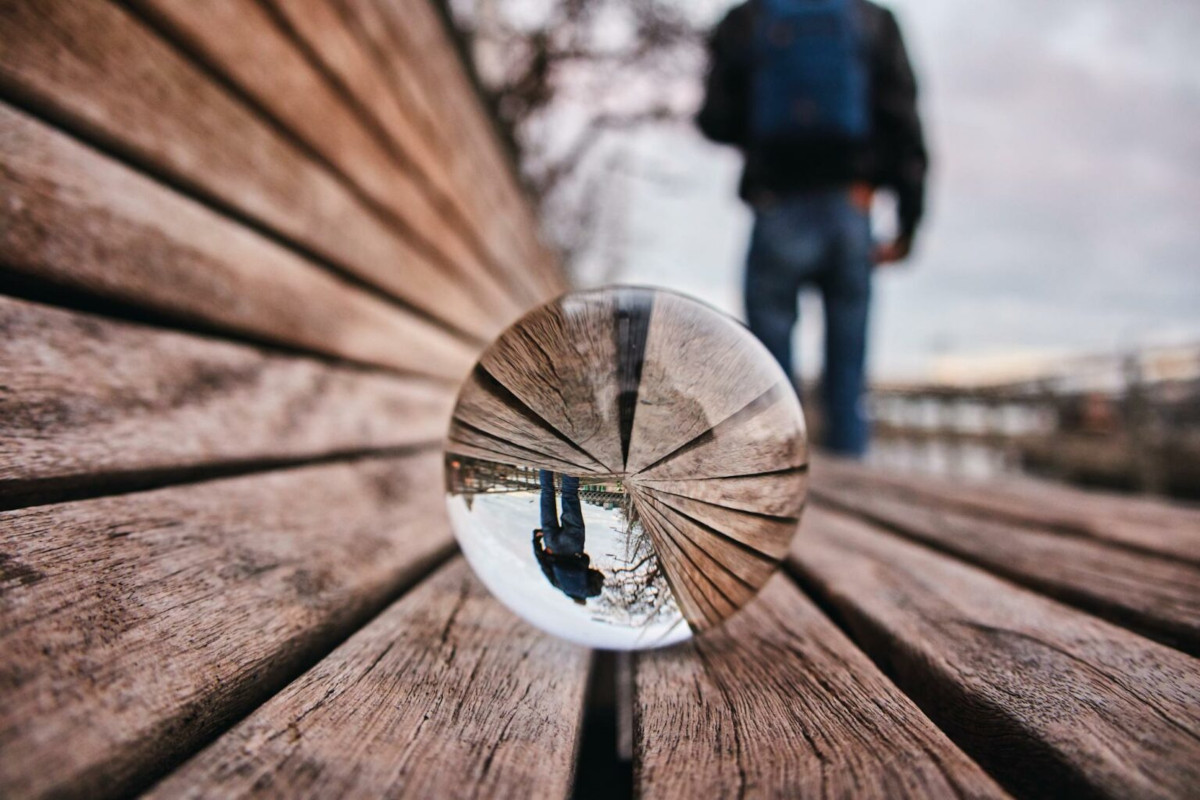 Photo of a person walking away from a glass ball on an outside bench. Their image is flipped upside down in the glass ball