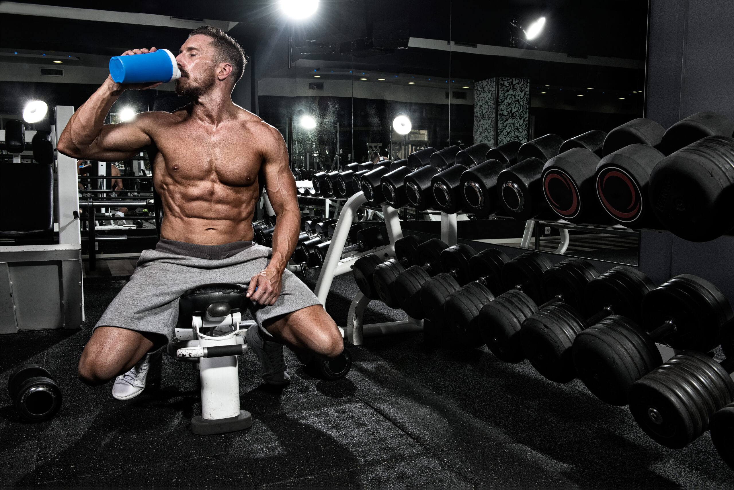 Muscled male bodybuilder sitting on the bench next to dumbbells weight stand and drinking protein shake smoothie. There are mirrors on the walls.