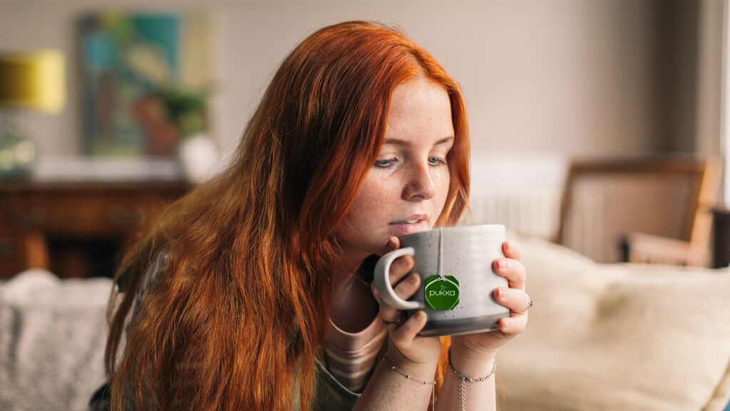 A young white woman with red hair holds a mug of tea with both hands.