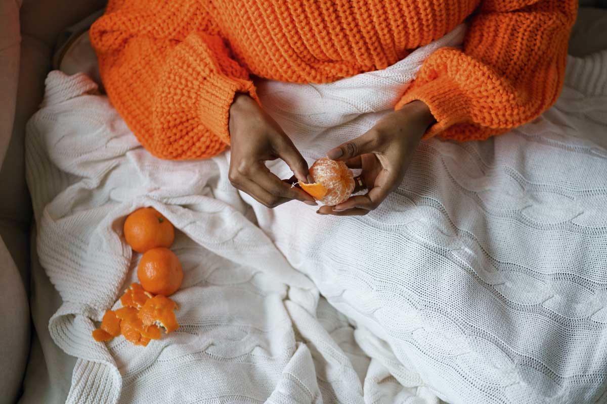 Afro Woman Hands Peeling Ripe Sweet Tangerine, Wear Orange Sweater, Lying In Bed Under The Blanket Person wearing orange jumper peeling an orange