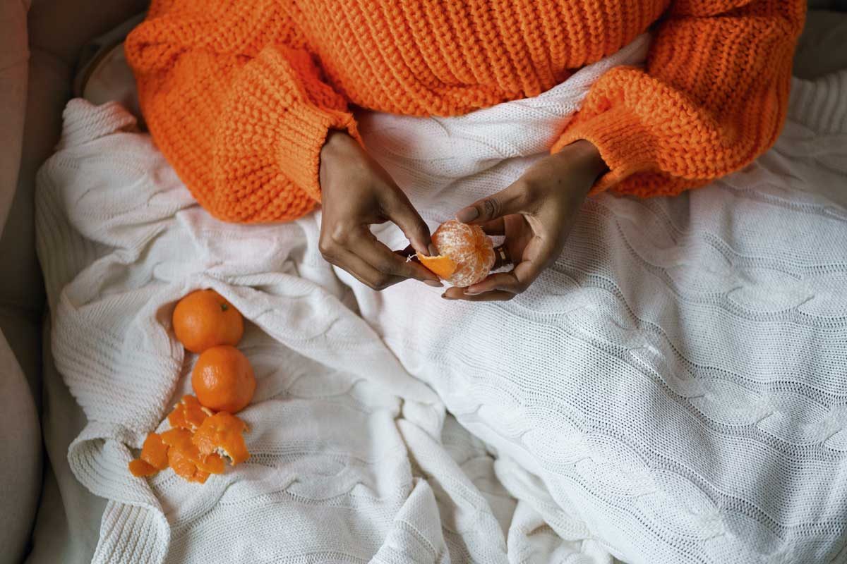 Afro Woman Hands Peeling Ripe Sweet Tangerine, Wear Orange Sweater, Lying In Bed Under The Blanket Person wearing orange jumper peeling an orange