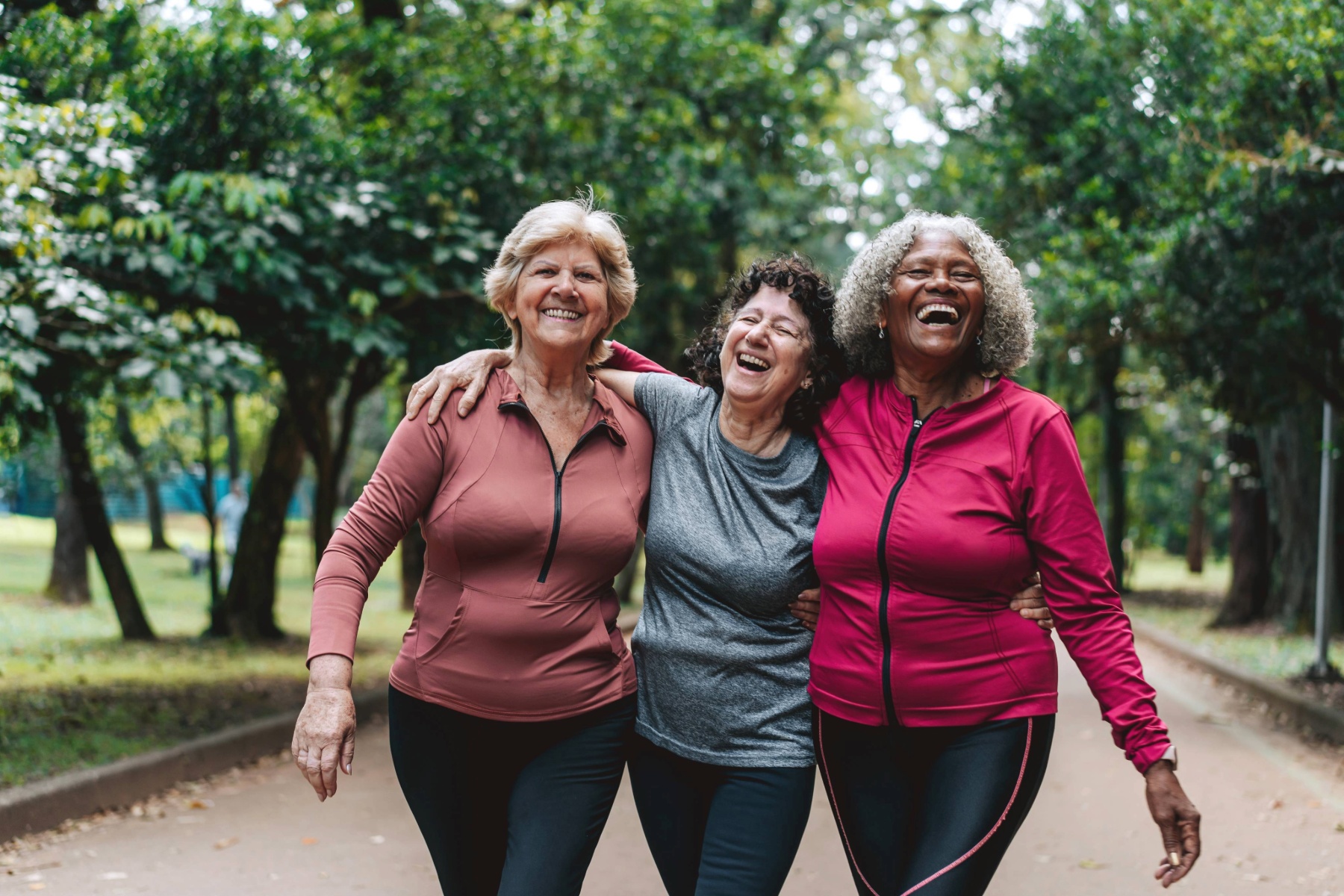 Three mature women on a walk in the park, smiling with their arms around each other