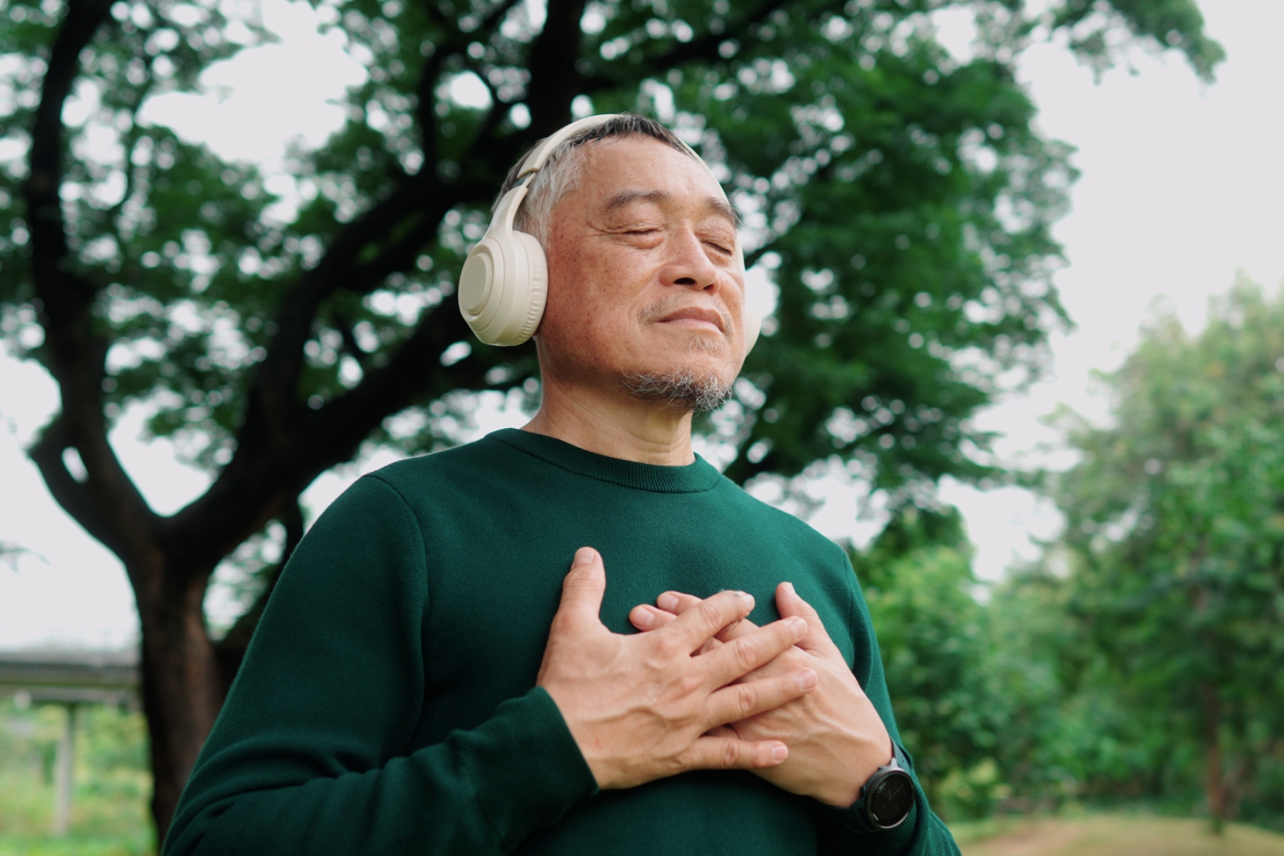 Older man in the park looking calm with eyes closed, headphones on and both hands on his chest