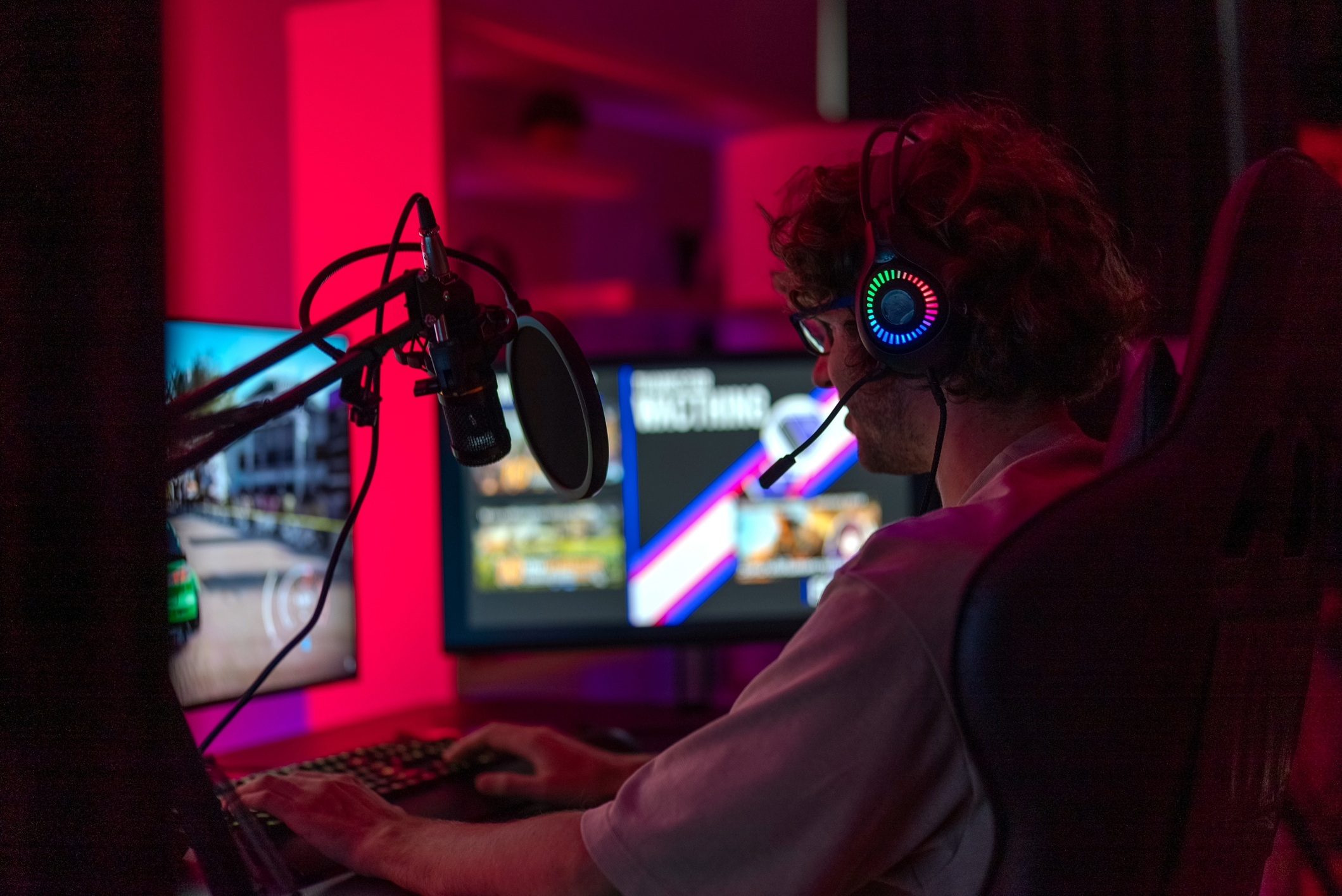 A person sitting in a darkened room, lit by the red glow of gaming lights, playing a video game on a monitor.