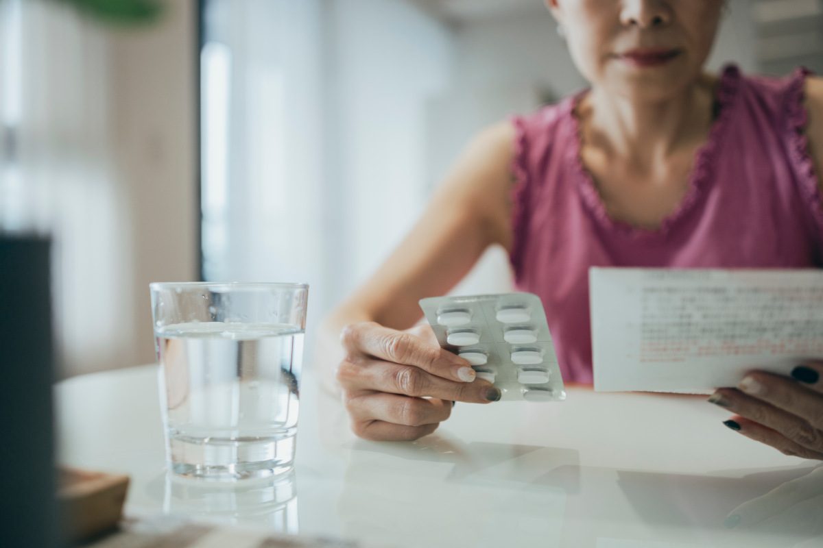 A person sat at a table holds pills in one hand and an information sheet about them in the other.