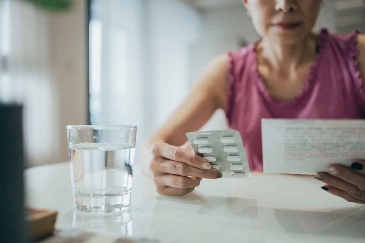 A person sat at a table holds pills in one hand and an information sheet about them in the other.