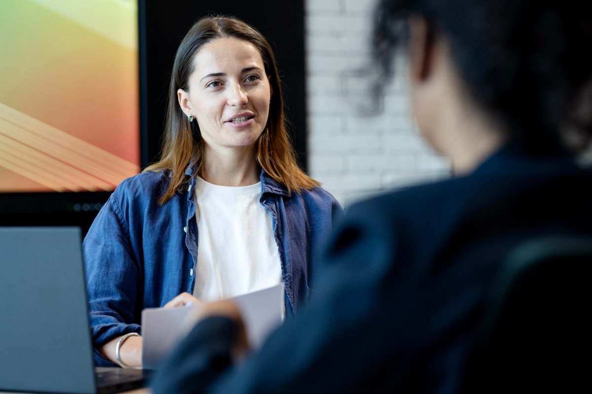 Photo of a person speaking to another blurred in the foreground. Interview setting