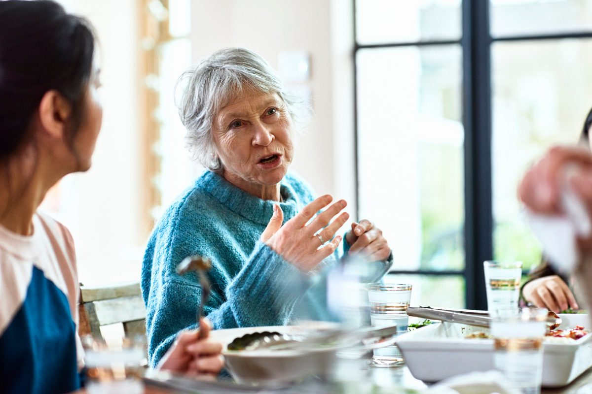 Candid portrait of woman in her 60s with grey hair, in conversation with friends, sitting at table with food and drink, woman listening in foreground