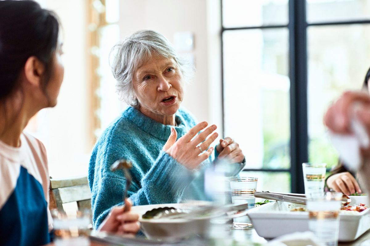 Candid portrait of woman in her 60s with grey hair, in conversation with friends, sitting at table with food and drink, woman listening in foreground