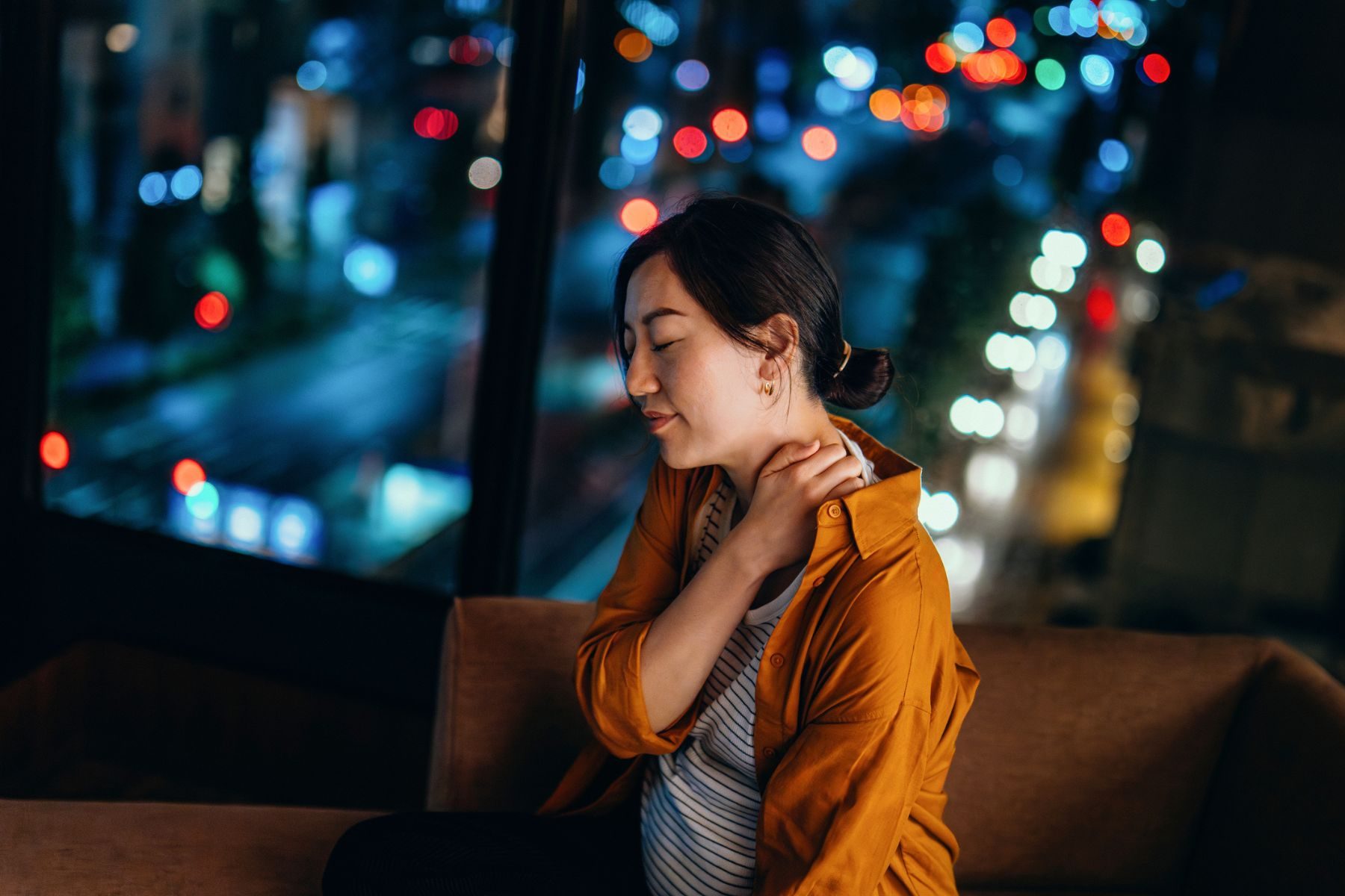 10 Expert-Approved Surprising Facts About Orgasms You Didn't Know 2 A woman sits in front of a window, rubbing her neck/shoulder in pain