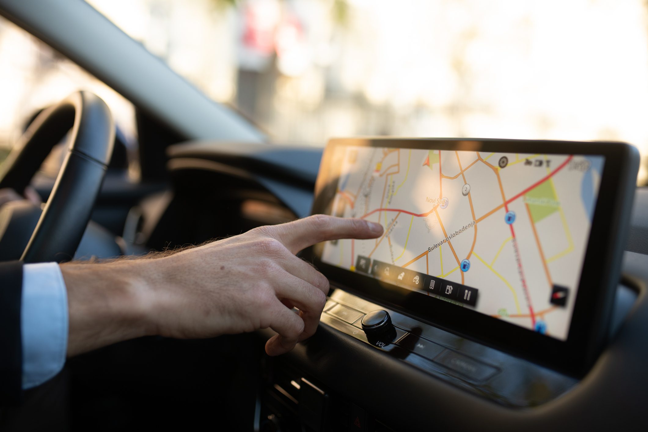 Close-up of a businessman’s hand adjusting the GPS on a modern car touchscreen. The man is sitting behind the wheel, wearing a suit, using the navigation system while driving, concept of business travel and technology.