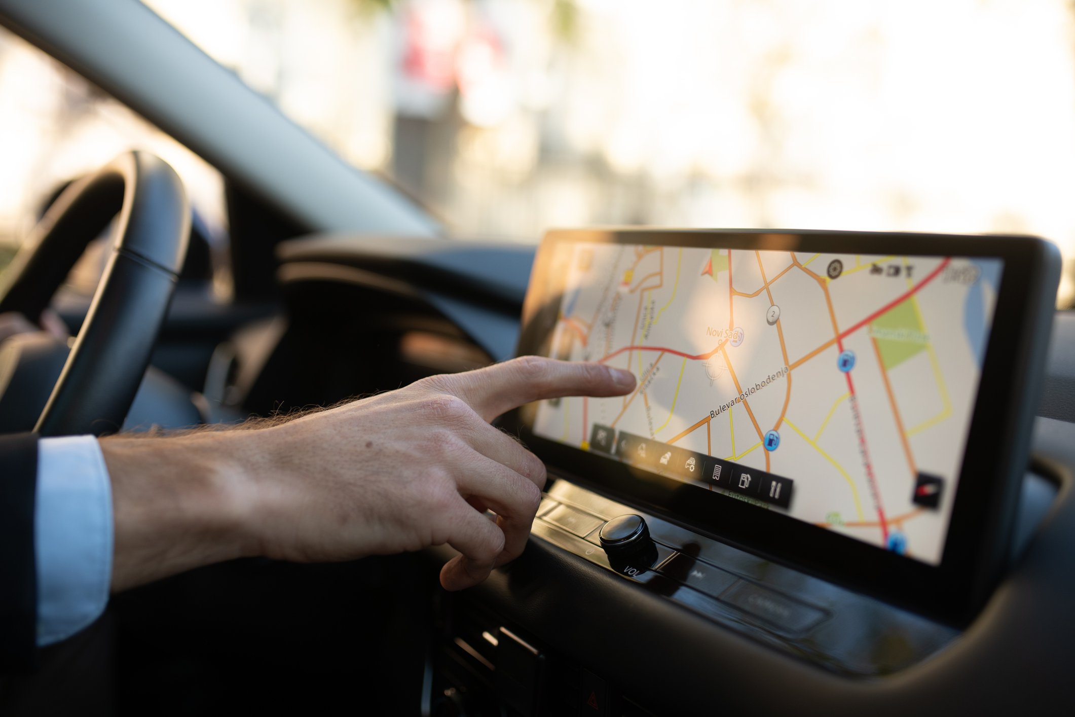 Close-up of a businessman’s hand adjusting the GPS on a modern car touchscreen. The man is sitting behind the wheel, wearing a suit, using the navigation system while driving, concept of business travel and technology.