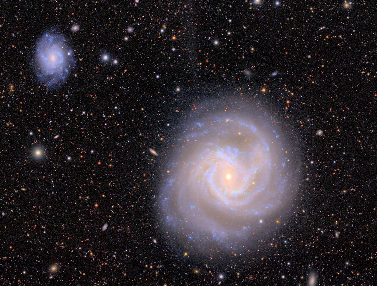 A purple spiral galaxy with a smaller one imaged in the top left corner of the image, surrounded by black starry sky.