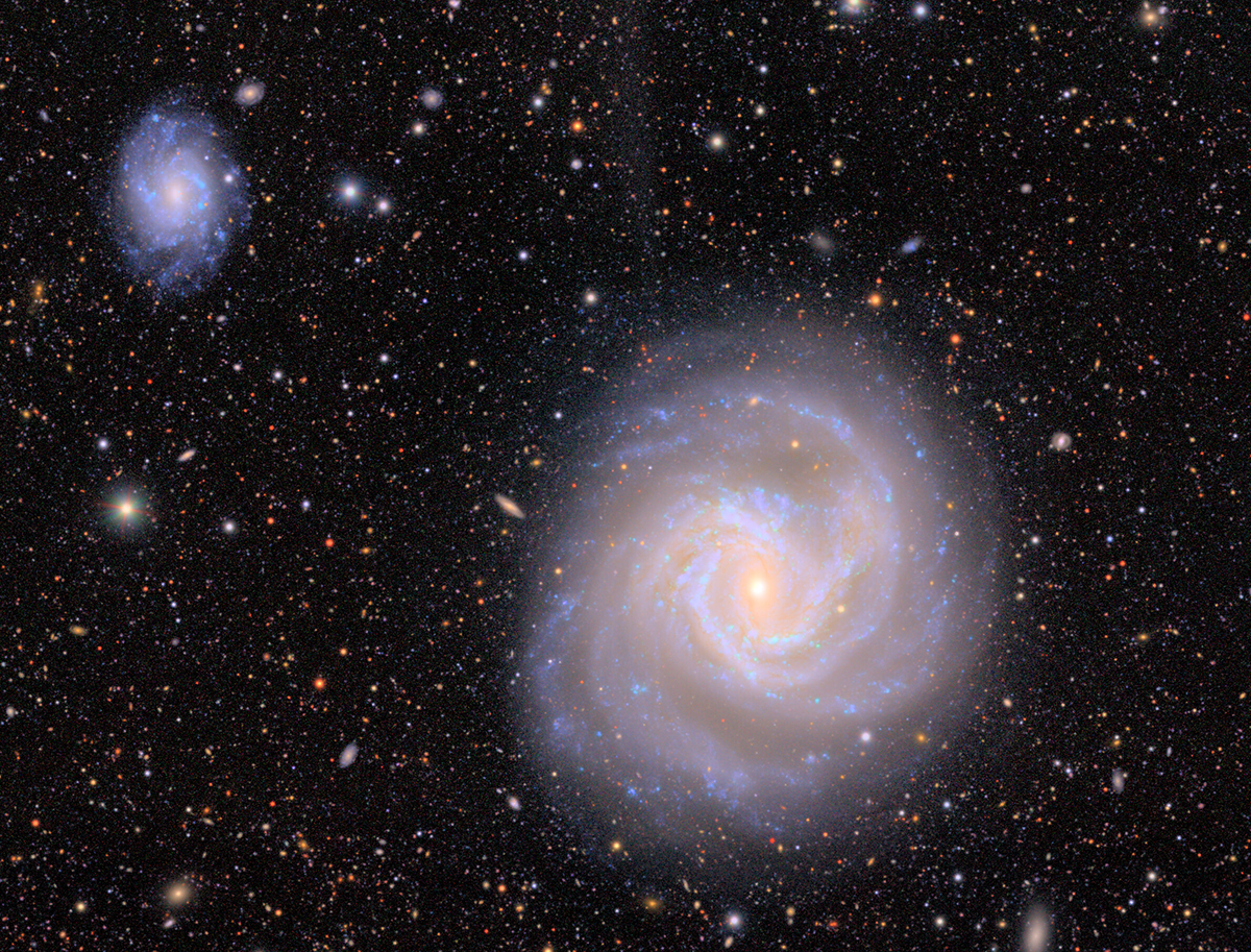 A purple spiral galaxy with a smaller one imaged in the top left corner of the image, surrounded by black starry sky.