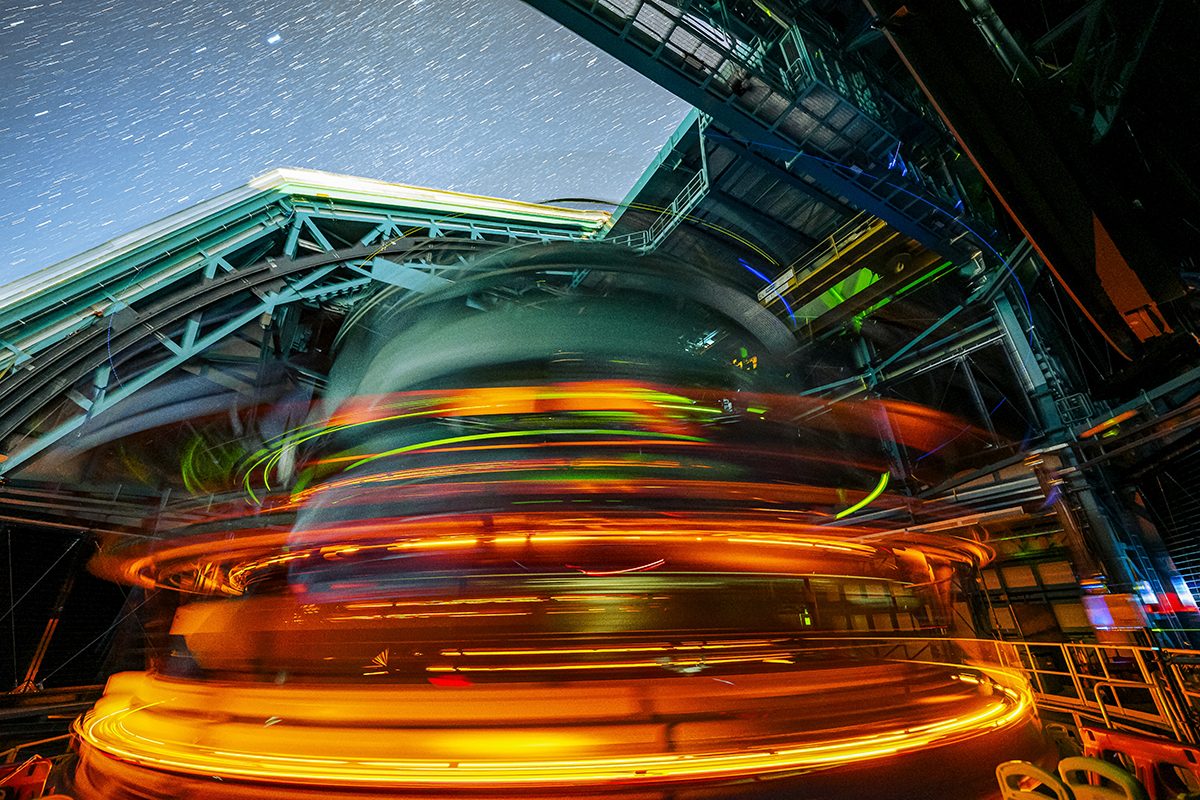 The oval shapped LSST camera in the vera rubin telescope is spinning, captured with a slow shutter, this leaves a trail of yellow and orange lights with the starry night sky seen above it.