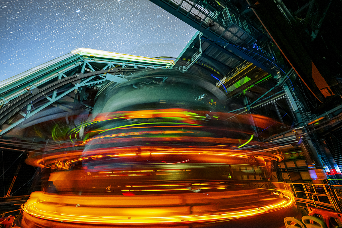 The oval shapped LSST camera in the vera rubin telescope is spinning, captured with a slow shutter, this leaves a trail of yellow and orange lights with the starry night sky seen above it.