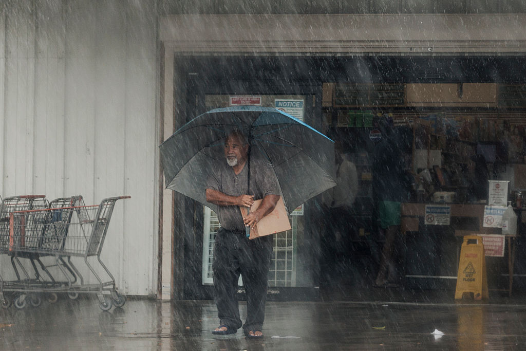 A man walks out of grocery store in torrential rain, carrying supplies and a large grey umbrella